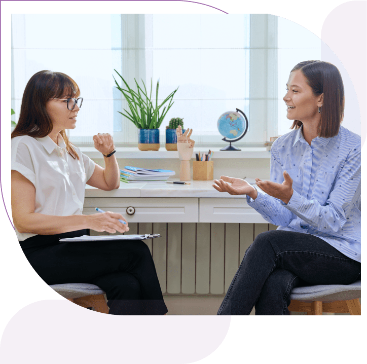 Two people engaged in conversation in an office setting, sitting on chairs facing each other. One is holding a clipboard. A desk with plants and a globe is in the background.